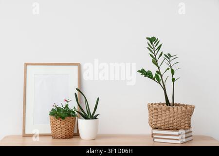 Grüne Pflanzen mit Büchern und leerem Rahmen auf dem Tisch in der Nähe der hellen Wand Stockfoto