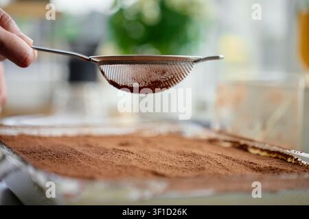 Hand hält Sieb mit Kakaopulver über Tiramisu-Dessert in Glasschale. Konzept für hausgemachtes italienisches Dessert und süßes Rezept Stockfoto