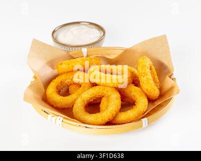 Knusprige goldene Calamariringe, serviert in einem geflochtenen Korb, der mit Pergamentpapier ausgelegt ist, neben einer kleinen Schüssel mit cremiger weißer Sauce. Helles, gleichmäßiges Licht hebt hervor Stockfoto