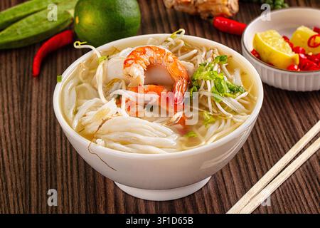 Nahaufnahme einer dampfenden Meeresfrüchte-Nudelsuppe mit prallen Garnelen, Reisnudeln, frischen Bohnensprossen. Garniert mit Limette, Chili, Kräutern auf Holztablett. W Stockfoto