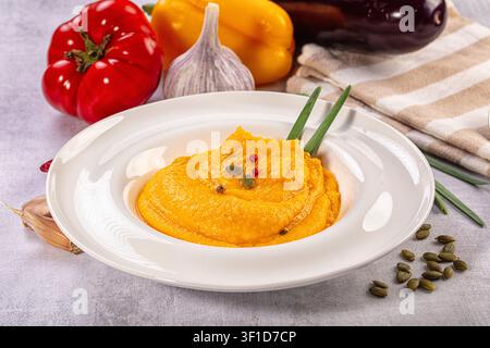 Leuchtend orangefarbene Kürbiskrem-Suppe serviert in einer weißen Keramikschale. Garniert mit grünen Zwiebelstielen und Kürbiskernen. Hintergrund zeigt rote Tomate gelb Stockfoto