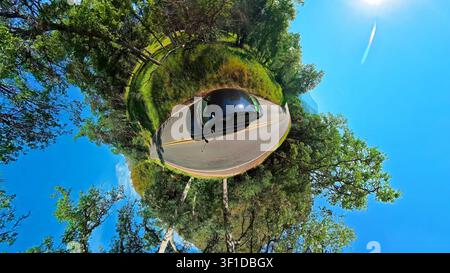 Tiny-Planet-Ansicht eines schwarzen Autos auf einer Waldstraße. Ein schwarzes Auto folgt einer kurvenreichen Waldstraße in einem lebhaften Tiny-Planet-Panorama unter einem strahlend blauen Himmel. Stockfoto