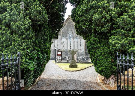 Lensborough Church of Ireland in Aghamore, County Longford, gesehen durch ein Eingangstor, das von dichten grünen Eiben und einem keltischen Kreuz eingerahmt ist. Stockfoto