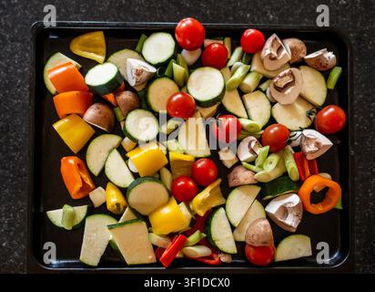 Eine Auswahl an frisch gehacktem rohem Gemüse, darunter Zucchini, Kirschtomaten, Paprika und Pilze, angeordnet auf einem schwarzen Backblech. Stockfoto