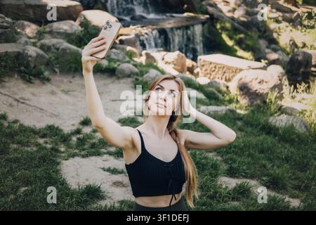 Frau macht Selfie mit Smartphone, während sie auf einer Matte im Waldpark sitzt. Content-Creator-Lebensstil, Social-Media-Influencer, Selfie-Fotografie-Trend. Stockfoto