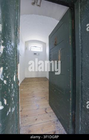 Kleine Zelle mit offener Metalltür, Holzfußboden und Überflutung von Licht aus einem Gitterfenster, Kilmainham-Gefängnis, Dublin, Irland Stockfoto