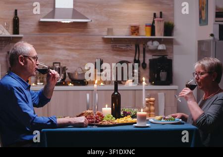 Älteres Paar stößt bei einem romantischen Abendessen in der Küche mit Weingläsern an und genießt gemeinsam eine Mahlzeit und die Feier zum Jahrestag. Stockfoto