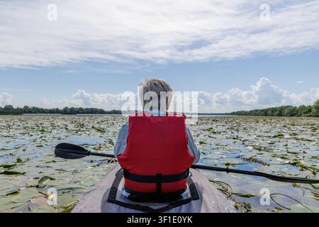 Eine Frau in einem Kajak mitten auf einem Fluss. Sie steht regungslos da und genießt die Schönheit. Das Wasser ist mit Blättern und Blumen von Seerosen bedeckt. Stockfoto