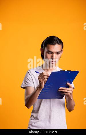 Ernsthafter männlicher Mensch hält ein Klemmbrett und überprüft tägliche Ziele und Aufgaben vor gelbem Hintergrund. Asiatisches männliches Model mit Stift in der Hand, liest Vertragsinformationen in einem modernen Studio. Stockfoto