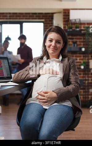 Schwangere Geschäftsfrau lächelt in die Kamera, während sie an einem Schreibtisch einer Finanzfirma sitzt, und präsentiert ein selbstbewusstes professionelles Büroporträt. Stockfoto