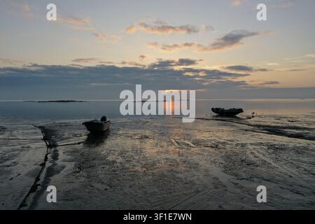 Zwei kleine Fischerboote liegen gestrandet auf einer glitzernden Schlickfläche, während ein goldener Sonnenaufgang den Horizont über der Taiwanstraße entflammt. Die Silhouetten der Boote ar Stockfoto