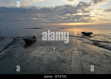 Zwei kleine Holzfischerboote liegen nebeneinander auf der freiliegenden Gezeitenwiese von Xiamen, während ein goldener Sonnenaufgang sie vor der leuchtenden Taiwanstraße silhouettiert Stockfoto