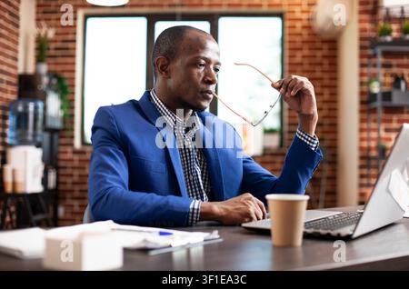 Afroamerikanischer Manager hält seine Brille, während er Firmenforschungsdaten auf einem Laptop in einem Startup-Büro mit konzentriertem Geschäftspapier studiert. Stockfoto