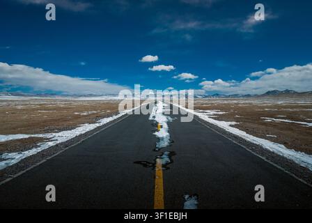 Gerade Asphaltstraße mit schmelzendem Schnee, die in den frühen Frühlingstagen auf die fernen Berge des Tibetischen Plateaus zuführt, Tibet Stockfoto