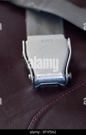 Eine Nahaufnahme, die sich auf eine zerkratzte silberne Metallschnalle mit Aufzugsschild-Text konzentriert, die gegen den dunkelroten Ledersitz eines Ai ruht Stockfoto