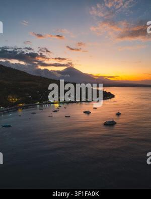 Luftaufnahme des Mount Agung, der über der Küste von Amed aufragt, mit Booten, die in der ruhigen See bei Sonnenuntergang in Amed, Bali, Indonesien, vor Anker liegen. Stockfoto