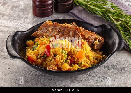 Geschmortes Lamm serviert über gewürztem Couscous mit roten Orangenpaprikaschoten. Präsentiert in schwarzer Keramikschale auf texturierter grauer Oberfläche. Frischer Rosmarin aus Holz Stockfoto