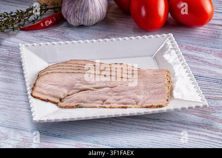 Geschnittenes Pastrami auf weißem Keramikteller mit gekräuseltem Rand angeordnet. Im Hintergrund sind Knoblauchknolle, rote Tomaten, Chili-Pfeffer und Thymian auf verwittertem Untergrund zu sehen Stockfoto