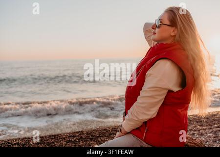 Strand-Sonnenuntergang-Frau sitzt auf Kiesufer in roter Weste und genießt den friedlichen Meerblick mit Kopierraum Stockfoto