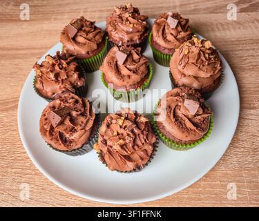 Eine Sammlung von frisch gebackenen Schokoladencupcakes, die mit cremiger Glasur, Schokoladenraspeln und Süßigkeitenstücken gekrönt sind, präsentiert auf einer weißen Platte Stockfoto