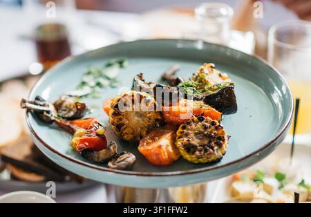 Ein farbenfrohes Gourmet-Gericht mit gegrilltem Mais, Pilzen, Kirschtomaten, Karotten und frischen Kräutern, serviert auf einem Keramikteller. Gesunde vegetarische Küche mit rustikaler Präsentation, selektivem Fokus und natürlichem Restaurantlicht. Stockfoto