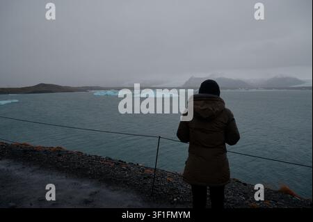 Einsame Gestalt in einer khakifarbenen Daunenjacke blickt über die Gewässer des Jökulsárlón, treibende blaue Eisberge und schneebedeckte Vulkanberge in der Ferne Stockfoto