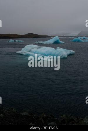 Leuchtende blaue Eisberge treiben über die dunklen, düsteren Gewässer der Gletscherlagune Jökulsárlón Stockfoto