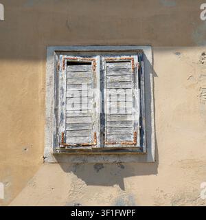 Piran, Slowenien. 26. Mai 2026. Ein verwittertes Holzfensterläden ist über einem in eine strukturierte Wand eingelassenen Fenster geschlossen. Der Fensterladen hat eine blasse, abblätternde Lackierung. Stockfoto