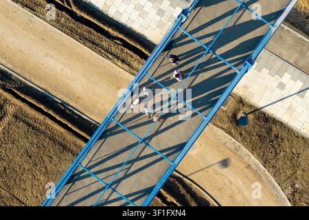Luftbild einer blauen Fußgängerbrücke mit Menschen, die darüber gehen und lange Schatten auf die umliegenden Erdwege und gepflasterten Gehwege in Belg werfen Stockfoto