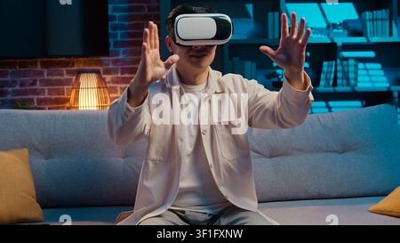 Asiatischer Mann mit moderner Technologie, der einen Virtual-Reality-Helm trägt, der abends im Wohnzimmer vr-Spiel spielt. Koreanisch Japanisch Chinesisch männlich genießt Stockfoto