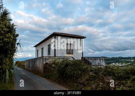 Ein weißes Haus mit einem roten Ziegeldach befindet sich hinter einer Betonmauer in Sao Miguel, Azoren, unter einem bewölkten Himmel. Stockfoto