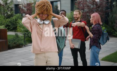 Enttäuschte afroamerikanische Schülerin, die Stress durch Mobbing von multikulturellen Schülern verspürt, die lachend grinsen in der Stadt, verärgerte weibliche Schülerin Stockfoto
