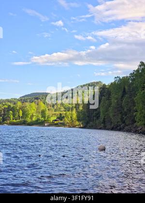 Friedliche Seelandschaft mit bewaldeten Hügeln, ruhigem Wasser und einem kleinen Haus unter einem klaren blauen Himmel. Stockfoto