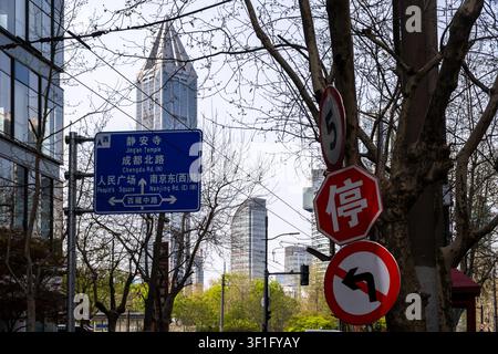 Wegweiser und Verkehrsschilder stehen zwischen Bäumen und Türmen in der Nähe des Jingan-Tempels in Shanghai, China, an einem Tag mit blauem Himmel Stockfoto