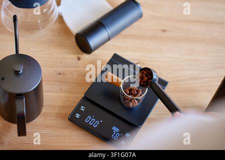 Geröstete Kaffeebohnen werden auf einer digitalen Waage mit manueller Kaffeemühle und Pour-Over-Kaffeearbeitsausrüstung auf einem Holztisch gemessen. Stockfoto
