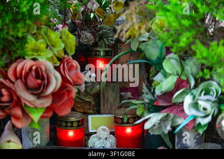 Leuchtend rote Gedenkkerzen auf einem Grab, das mit künstlichen Rosen, einer kleinen Engelsstatue und einem leeren Namensschild geschmückt ist Stockfoto
