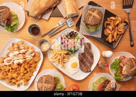 "Überkopf-Festmahl aus Sesamburgern, Steak-Kombi-Teller, beladenen Pommes und Sandwiches auf Holztable" Stockfoto