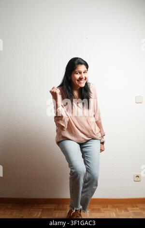 Junge Frau lächelt herzlich vor einer schlichten, strukturierten Wand. Sie trägt einen weichrosa Pullover und verwaschene Jeans. Ihr dunkles Haar fällt locker über die Schultern. Braun Stockfoto