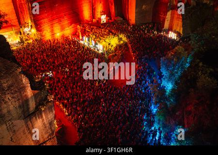 Luftaufnahme einer großen Menschenmenge, die ein Open-Air-Musikfestival in einem tiefen Steinbruch besucht, beleuchtet von lebendigem rotem und blauem Bühnenlicht Stockfoto