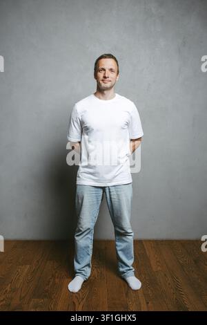 Ein entspannter Mann, der ein weißes T-Shirt und helle Jeans trägt, steht barfuß auf einem Holzboden vor einer neutralen grauen Wand, lächelt subtil und schaut direkt Stockfoto