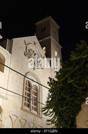 Fassade der Kathedrale von Ostuni mit ihrem dekorativen Rosettenfenster und Glockenturm, die von warmem Licht gegen den dunklen Nachthimmel beleuchtet werden. Italien, Apulien, Giovinazzo Stockfoto