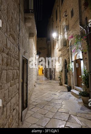 Schmale Kopfsteinpflasterstraße schlängelt sich durch ein historisches Stadtzentrum bei Nacht, beleuchtet von Straßenlaternen und Umgebungslicht. Italien, Apulien, Giovinazzo, 19. Jul Stockfoto