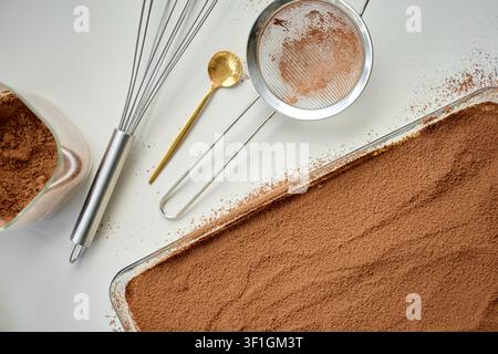 Tiramisu-Dessert mit Kakaopulver bedeckt in der Backform, mit Sieb und Kakaoschüssel auf dem Tisch. Konzept für hausgemachtes Dessert und italienische Süßspeise Stockfoto