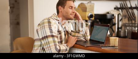 Nahaufnahme Porträt eines gutaussehenden Mannes, der an der Theke sitzt und mit einem fröhlichen Lächeln auf den Laptop schaut, Kaffee trinkt, im Internet surft, wattiert Stockfoto