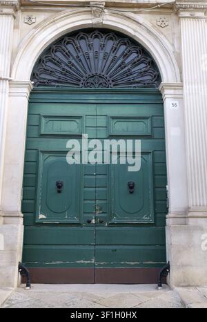Verzierte grüne Holztür mit dunklen Löwenkopf-Klöpfern und einem kunstvollen schmiedeeisernen Oberlicht. Italien, Apulien, Molfetta, 21. Juli 2025 Stockfoto