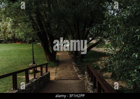 Eine kleine Holzbrücke führt zu einem schattigen Pfad unter alten Bäumen. Die friedliche Umgebung bietet eine ruhige Oase, umgeben von üppiger Vegetation. Stockfoto