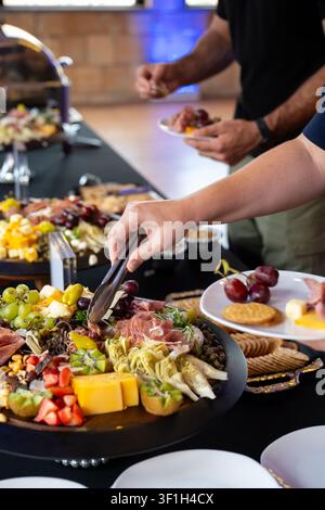 Gäste verwenden Servierzangen, um Käse, Obst, gepökeltes Fleisch, Cracker und Gemüse von einem Charcuterie-Tisch bei einer Indoor-Veranstaltung auszuwählen. Stockfoto