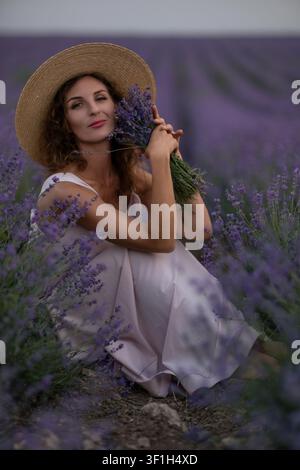Junge Frau mit Strohhut sitzt in einem blühenden Lavendelfeld und hält frische violette Blumen bei Sonnenuntergang Stockfoto