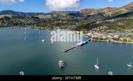 Luftaufnahme des Hafens von Akaroa mit Booten, die in der Bucht vor Anker liegen, einem langen Pier, der sich ins Wasser erstreckt, und der Stadt, die sich an sanfte grüne Hügel in A schmiegt Stockfoto