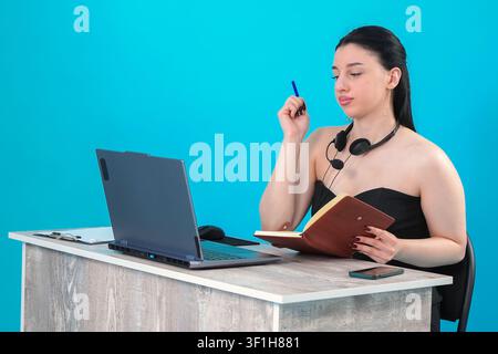 Konzentrierte junge Geschäftsfrau schreibt in ein Notizbuch, während sie einen Laptop benutzt Stockfoto